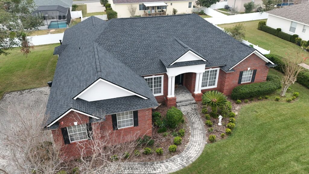 Aerial view of a single-story brick house with a black shingle roof, white trim, columns at the entrance, neatly trimmed bushes, and a curved walkway surrounded by a well-maintained lawn and landscaping.
