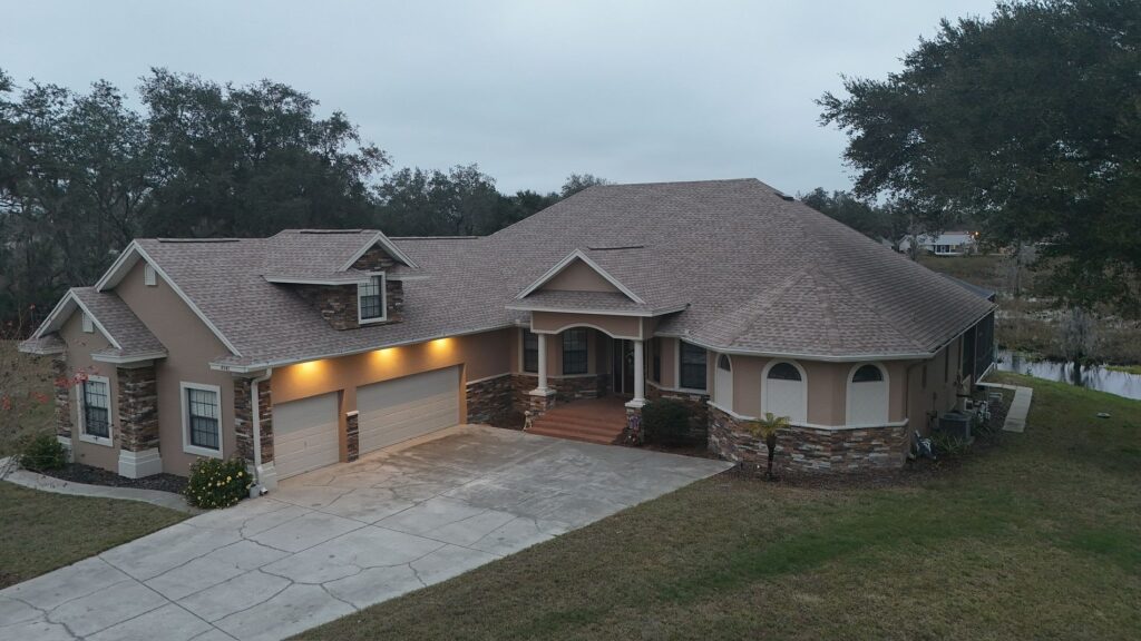 A large single-story house with stone and tan siding, a three-car garage, outdoor lights on, and a curved driveway. The yard has grass, a few trees, and a pond is visible in the background.