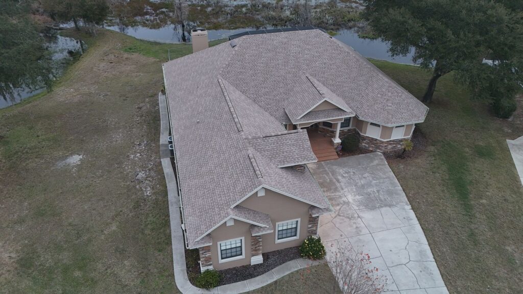 Aerial view of a single-story beige house with a gray shingle roof, surrounded by grass, trees, and a small pond in the background. A driveway leads up to the front entrance.