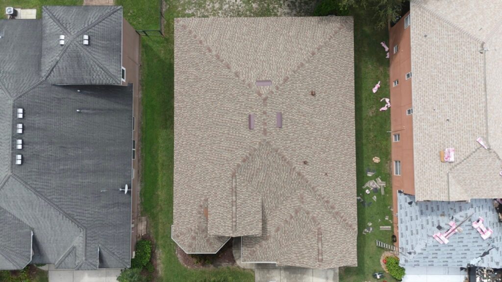 Aerial view of three adjacent houses, focusing on the roofs. The middle roof has a brownish pattern and three vents. The left and right roofs are darker, with the right one partially covered in construction materials.