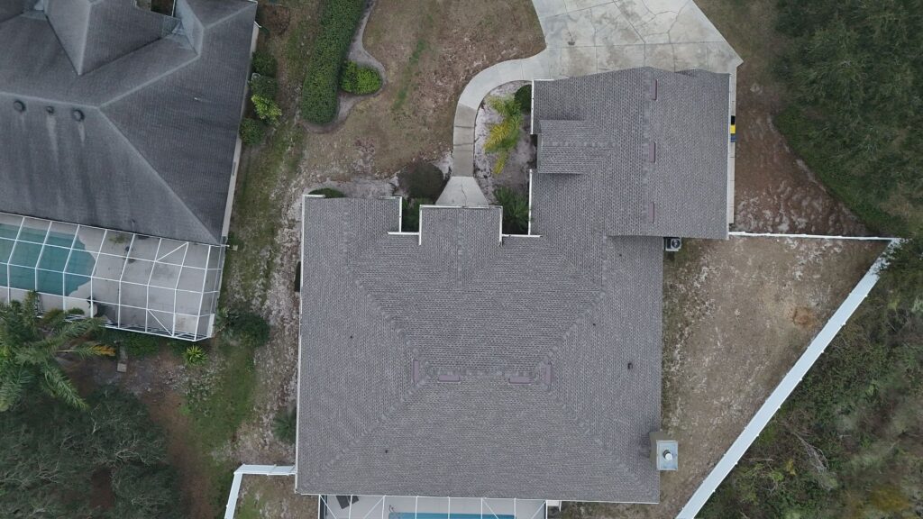 Aerial view of a house with a gray roof, surrounded by a driveway, yard, white fence, and neighboring homes; one neighbor has a screened pool area.
