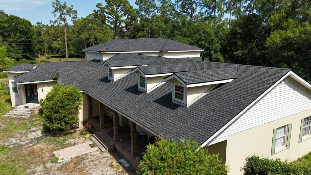 A beige house with a dark shingled roof, multiple dormer windows, and a covered front porch is surrounded by green trees and a partially grassy, partially sandy yard.