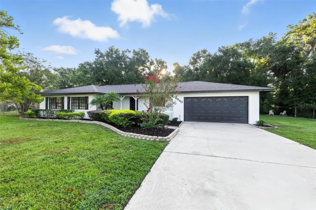 Single-story white house with dark gray shutters and a matching double garage. A large driveway leads to the house, with well-kept green lawn, shrubs, and trees surrounding the property under a blue sky.
