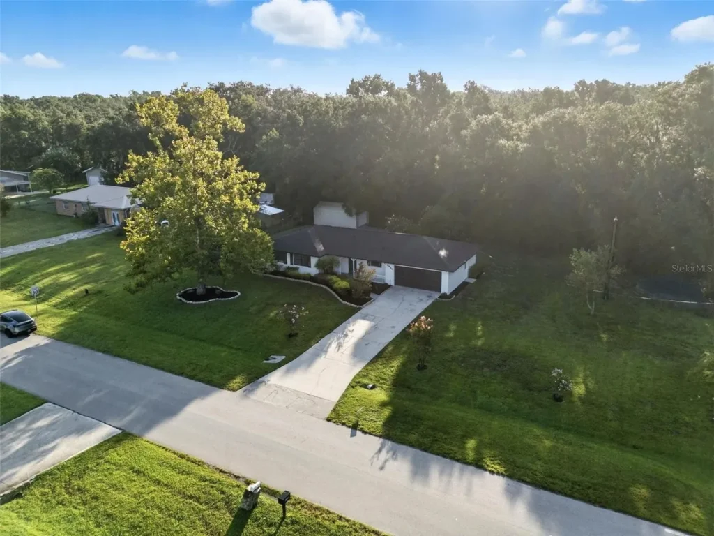 Aerial view of a single-story house with a dark roof, wide driveway, and well-kept green lawn, surrounded by trees and neighboring homes under a bright blue sky with scattered clouds.