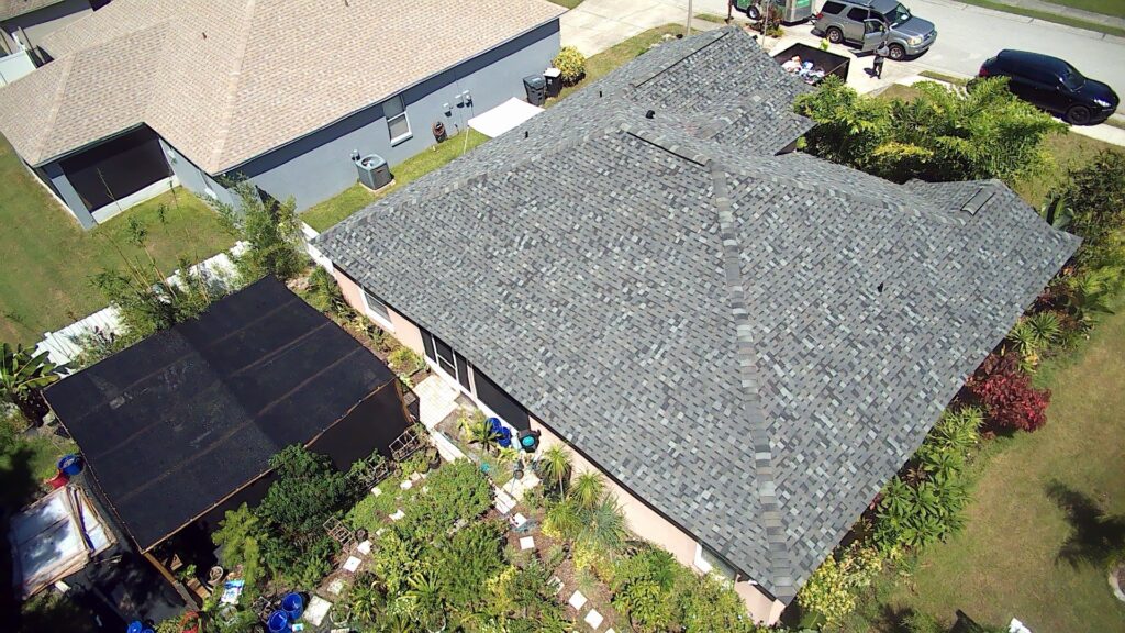 Aerial view of a house with a gray shingle roof, a fenced backyard garden, and a black screened structure. Nearby houses, a driveway, and parked vehicles are also visible.