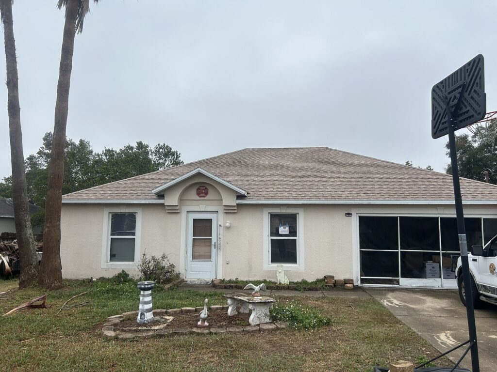 A single-story house with a light-colored exterior, a central front door, two windows, and a screened garage. The yard has birdbaths, statues, and a basketball hoop on the right. Sky is overcast.