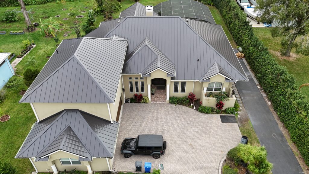 Aerial view of a large beige house with a gray metal roof, paver driveway, black SUV parked in front, surrounding lawn, and tall green hedges bordering the property.