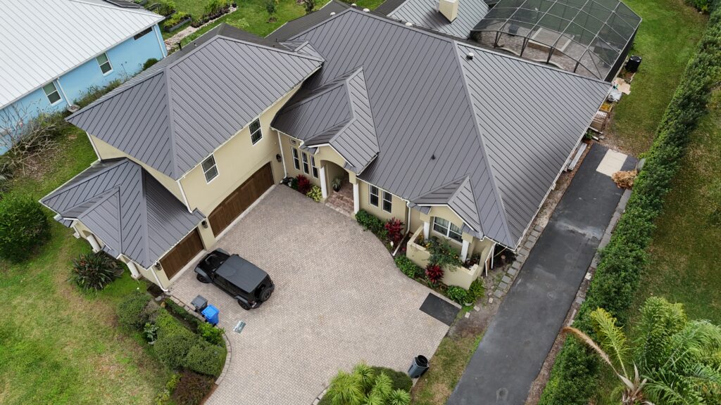 Aerial view of a modern beige house with a gray metal roof, double garage, black car parked on a paved driveway, and lush green landscaping surrounding the property.