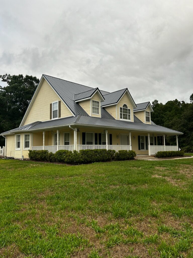 A large, two-story yellow house with a gray metal roof, white trim, and a wide wraparound porch sits on a green lawn under a cloudy sky. Bushes line the front of the porch. Trees are visible in the background.