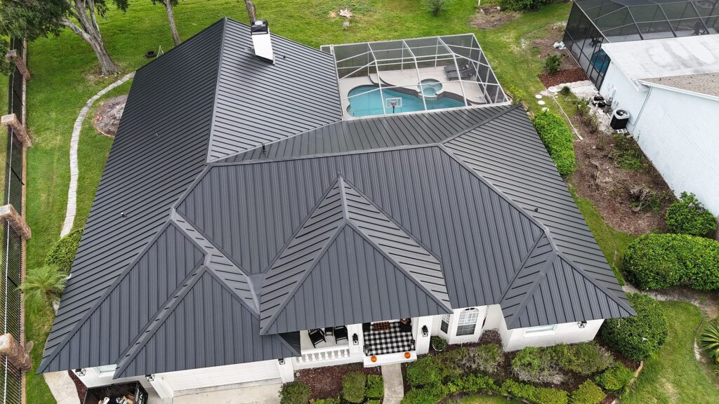 Aerial view of a house with a dark metal roof, surrounded by green grass and shrubs. In the backyard, there is a screened enclosure over a swimming pool and patio area.