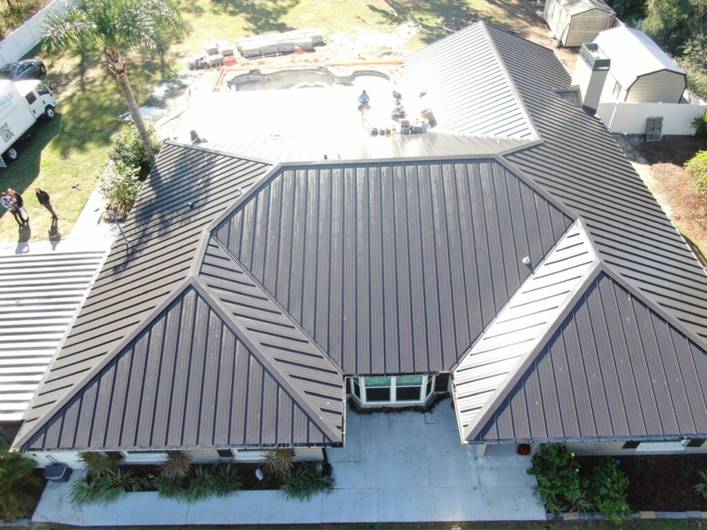 Aerial view of a house with a dark metal roof, surrounded by greenery. In the backyard, people are working near an unfinished swimming pool. There are sheds and vehicles visible on the property.