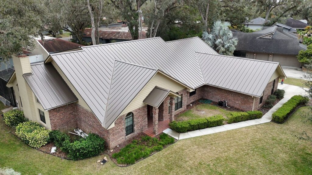 Aerial view of a single-story brick house with a light brown metal roof, surrounded by grass, shrubs, and trees. A concrete walkway borders the front and side of the house. Neighboring houses are visible in the background.