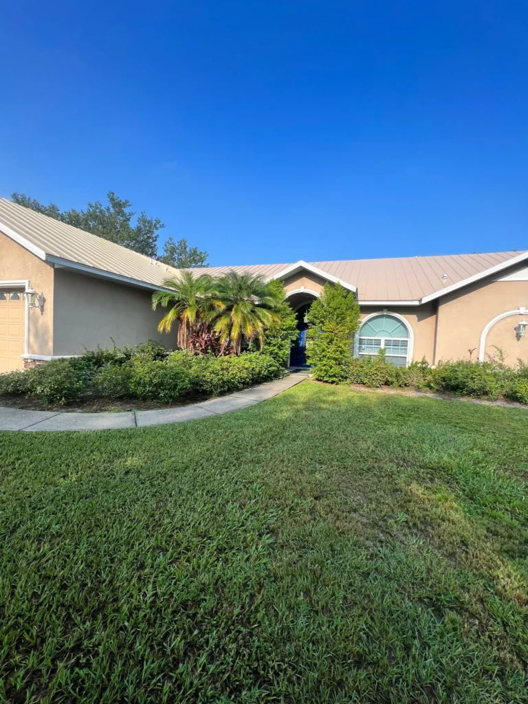 A single-story house with a light-colored exterior, metal roof, and arched doorway. There are palm trees, shrubs, and a neatly cut lawn under a clear blue sky.