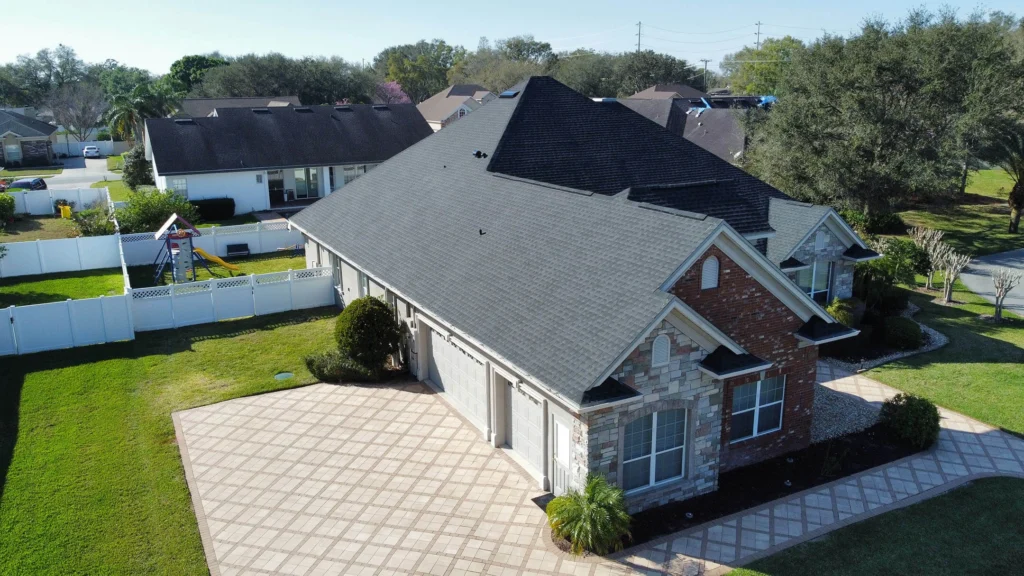 Aerial view of a suburban house with a dark roof, brick and stone facade, and a large tiled driveway. The backyard features a white fence and a children's playset. Other houses and trees are visible in the background.