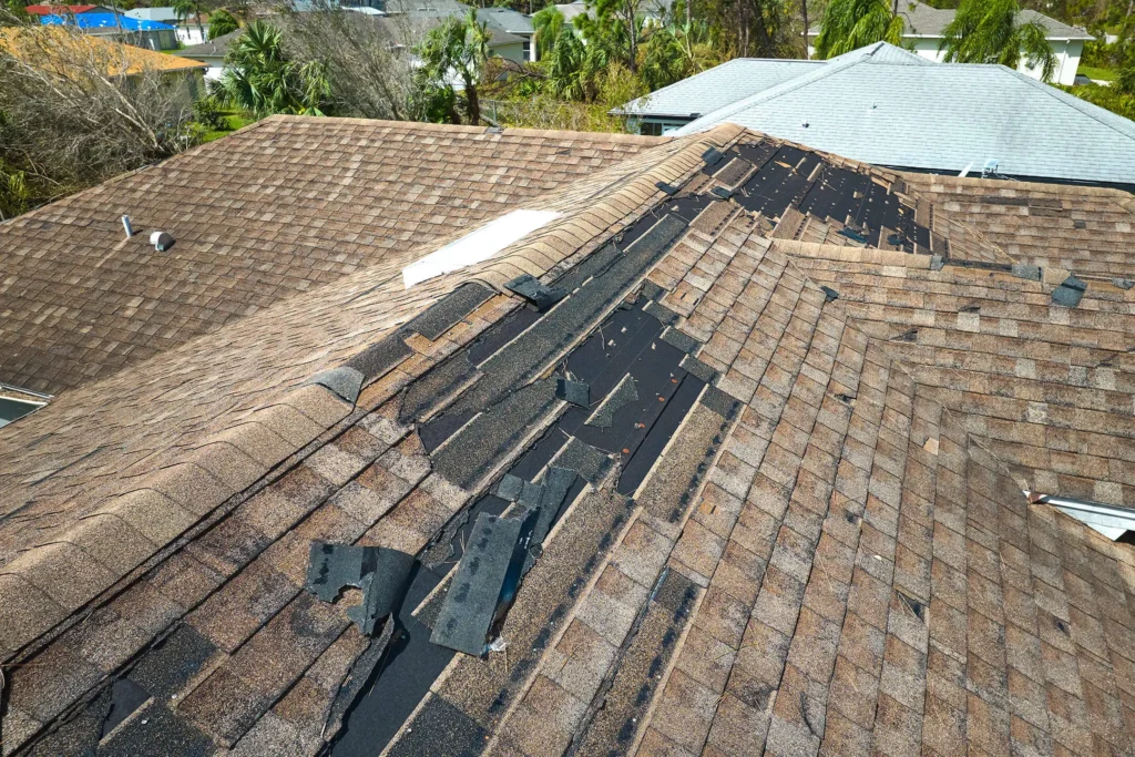 A house roof with numerous missing and damaged shingles, exposing black underlayment beneath. The roof damage runs along the ridge, indicating possible storm or wind damage. Nearby houses with intact roofs are visible.