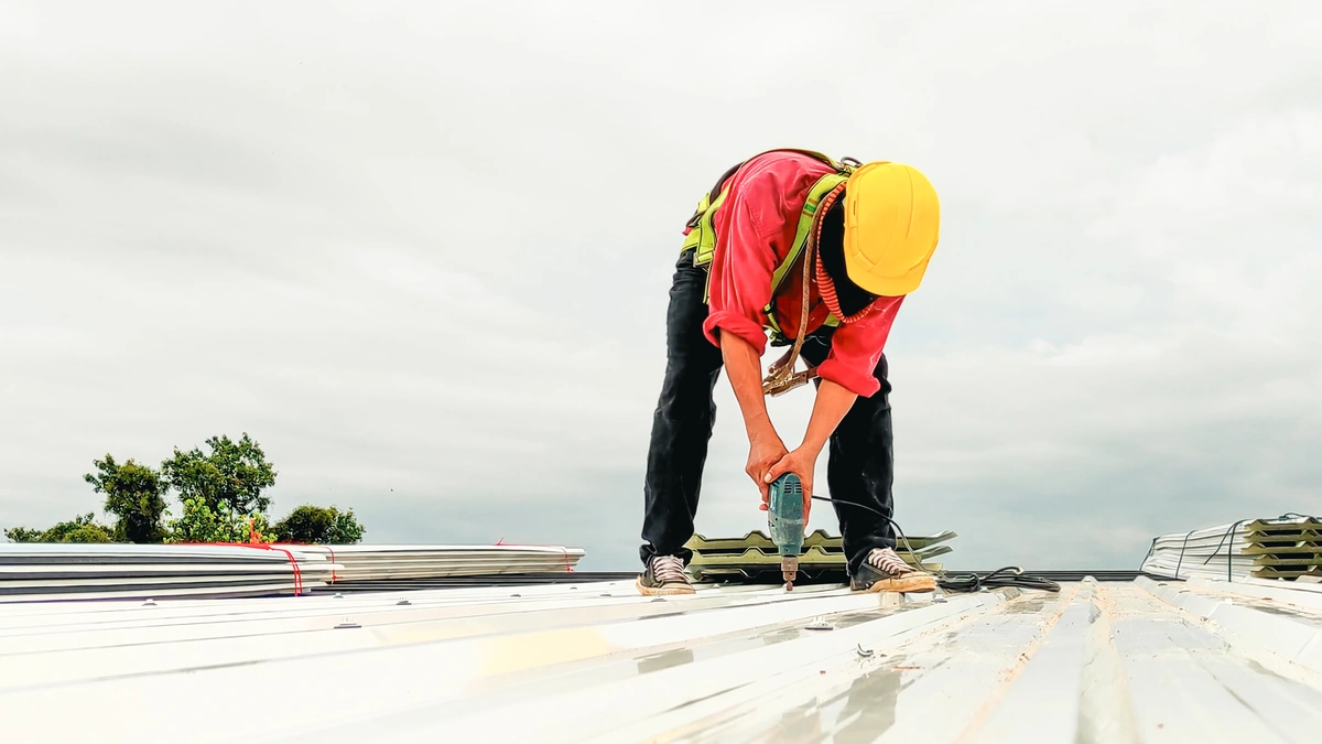 A construction worker in a yellow hard hat and red shirt uses a power drill to attach metal sheets during a commercial roof replacement under a cloudy sky.