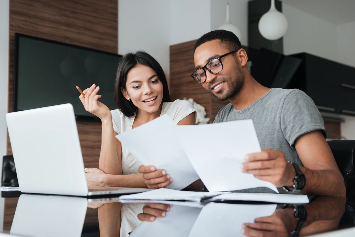 A smiling man and woman sit at a table with a laptop, reviewing roof financing documents together in a modern home setting. The woman holds a pencil, and both appear engaged and focused on the papers.