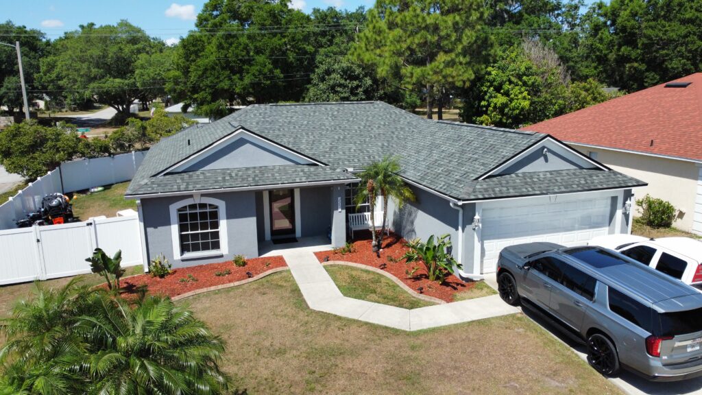 A gray single-story house with a neatly landscaped front yard, red mulch around the plants, a white fence, and two vehicles parked in the driveway. Trees and neighboring houses are visible in the background.