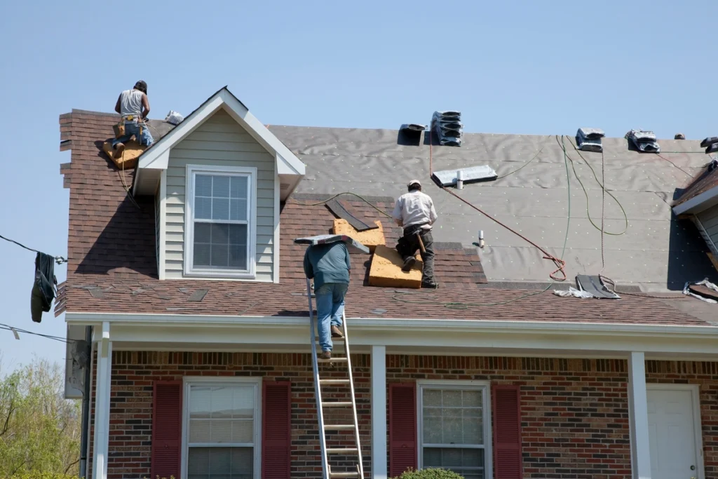 Three workers are repairing the roof of a brick house using ladders and tools. Roofing materials, shingles, and supplies for a roof overlay are scattered around as they work under a clear sky.