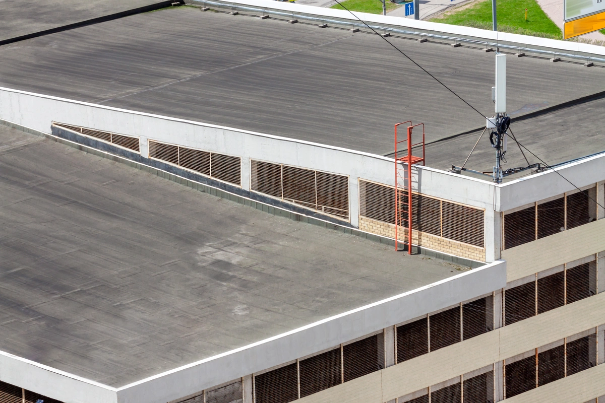 A flat industrial building roof—one of the common commercial roof types—features white walls, horizontal vents, a red metal ladder at the edge, and electrical cables connected to equipment on top.