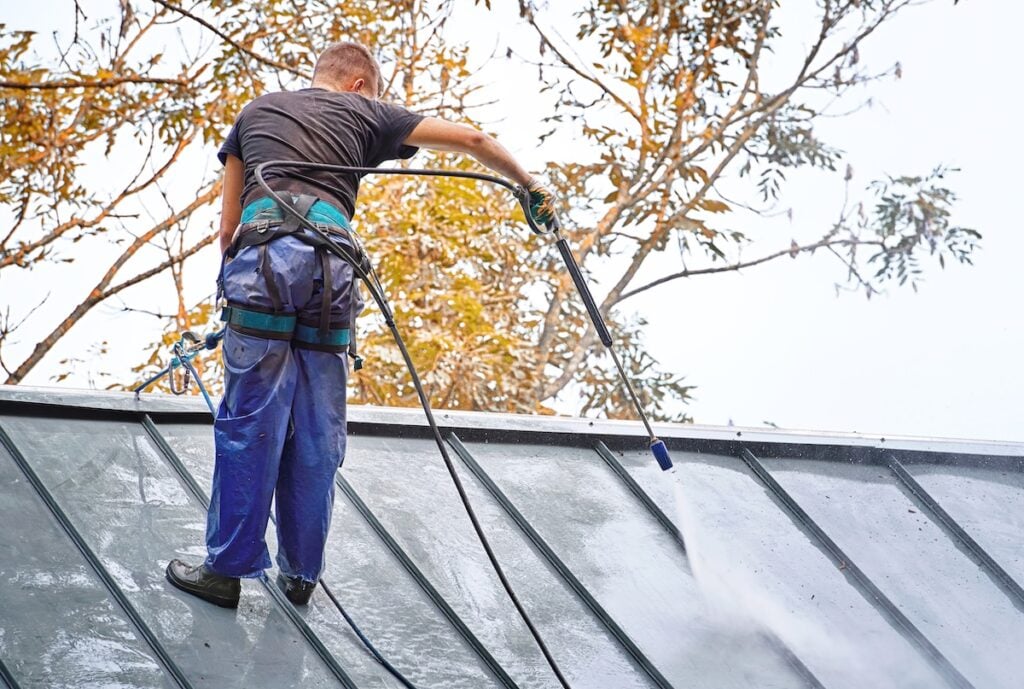 Man, cleaner with high pressure water sprayer wash steel dirty roof.Worker in waterproof uniform with pressure washer gun wash roof of private house. Roof cleaning with water pressure cleaner