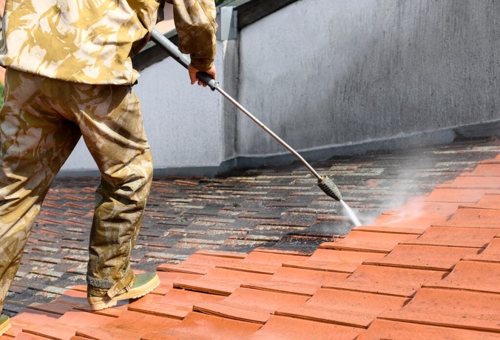 Professional in uniform using high pressure machine to clean rooftop from dirt and lichen. Before and after situation. Clean and dirty roof tiles.