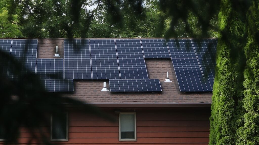 Solar photovoltaic panels on the roof of a red house framed by a foreground of green trees. Solar Panel Module technology creating renewable energy in