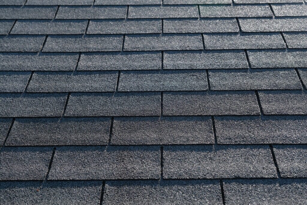 Close-up view of a roof covered with overlapping rectangular composition shingles, arranged in a neat, uniform pattern. The shingles have a rough, textured surface in varying shades of gray.