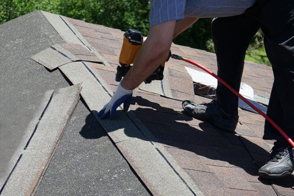A person wearing gloves uses a nail gun to attach shingles to a roof on a sunny day, with greenery visible in the background. should i stay home during a roof replacement