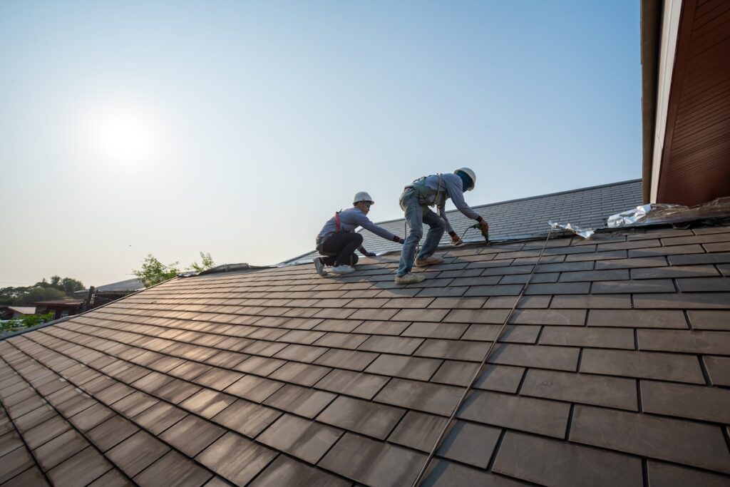 Two construction workers wearing safety gear are installing or repairing roof shingles on a sloped house roof under a clear sky with the sun shining. should i stay home during a roof replacement