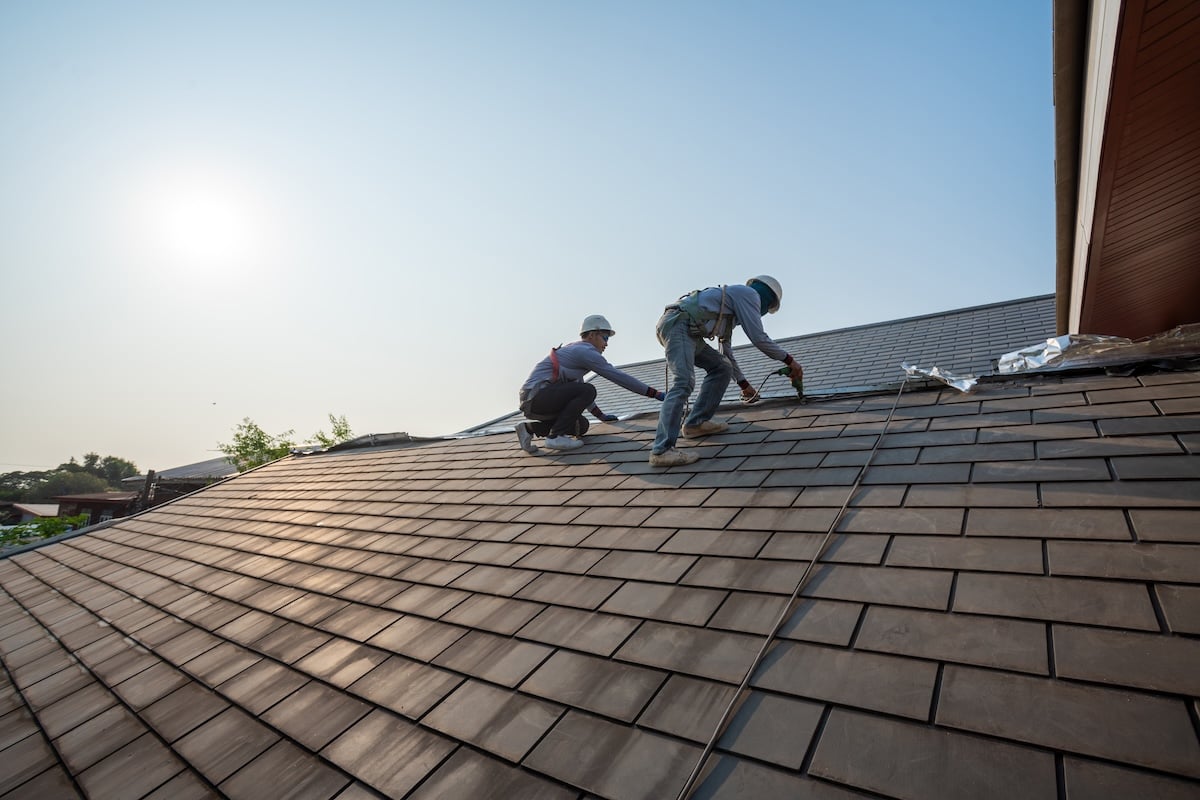 Two construction workers wearing safety gear are installing or repairing roof shingles on a sloped house roof under a clear sky with the sun shining. should i stay home during a roof replacement