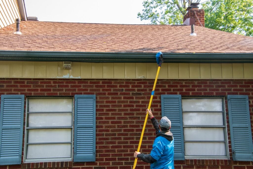 A person uses a long yellow pole with a brush to clean the gutters of a brick house with blue shutters and a brown roof, showing the care and attention often seen in soft wash roof cleaning. soft wash roof cleaning