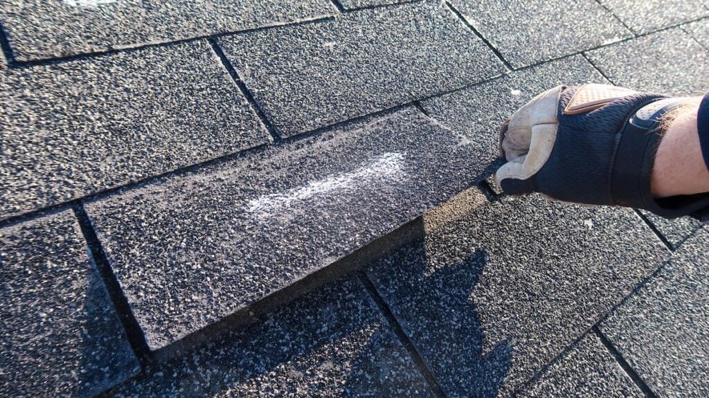 A gloved hand lifts a shingle on an asphalt shingle roof, revealing white chalk marking on the shingle, possibly indicating inspection or repair work.