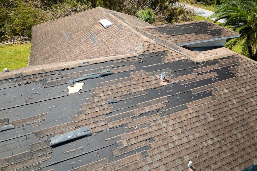 A house roof with many missing and damaged shingles, exposing the black underlayment beneath. Debris is scattered on the roof, indicating recent storm or wind damage.