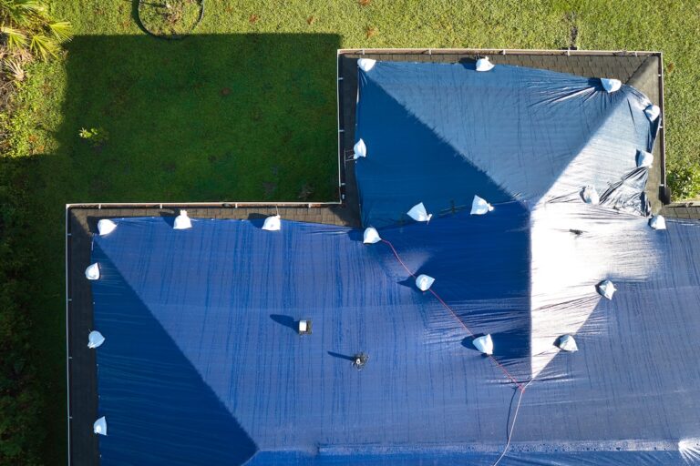 Aerial view of a house roof covered with large blue tarps secured by white sandbags, surrounded by green grass and partial tree shadows.