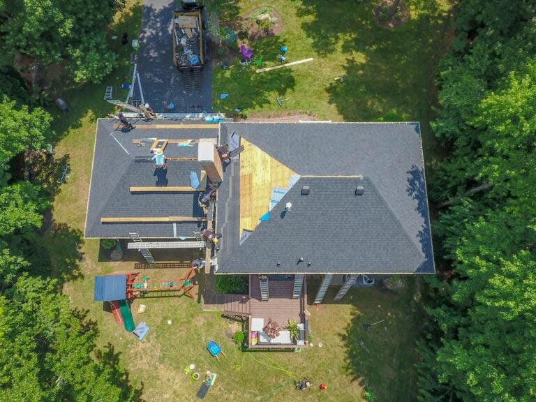 Aerial view of a house with workers repairing the roof; some shingles have been removed, exposing wooden panels. Construction materials and equipment are scattered on the lawn and driveway, surrounded by trees.