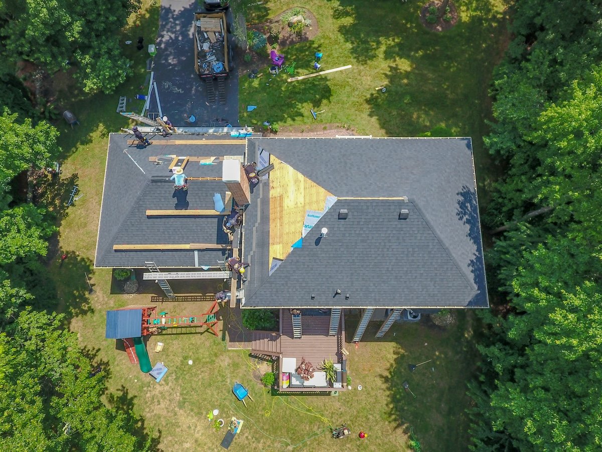 Aerial view of a house with workers repairing the roof; some shingles have been removed, exposing wooden panels. Construction materials and equipment are scattered on the lawn and driveway, surrounded by trees.