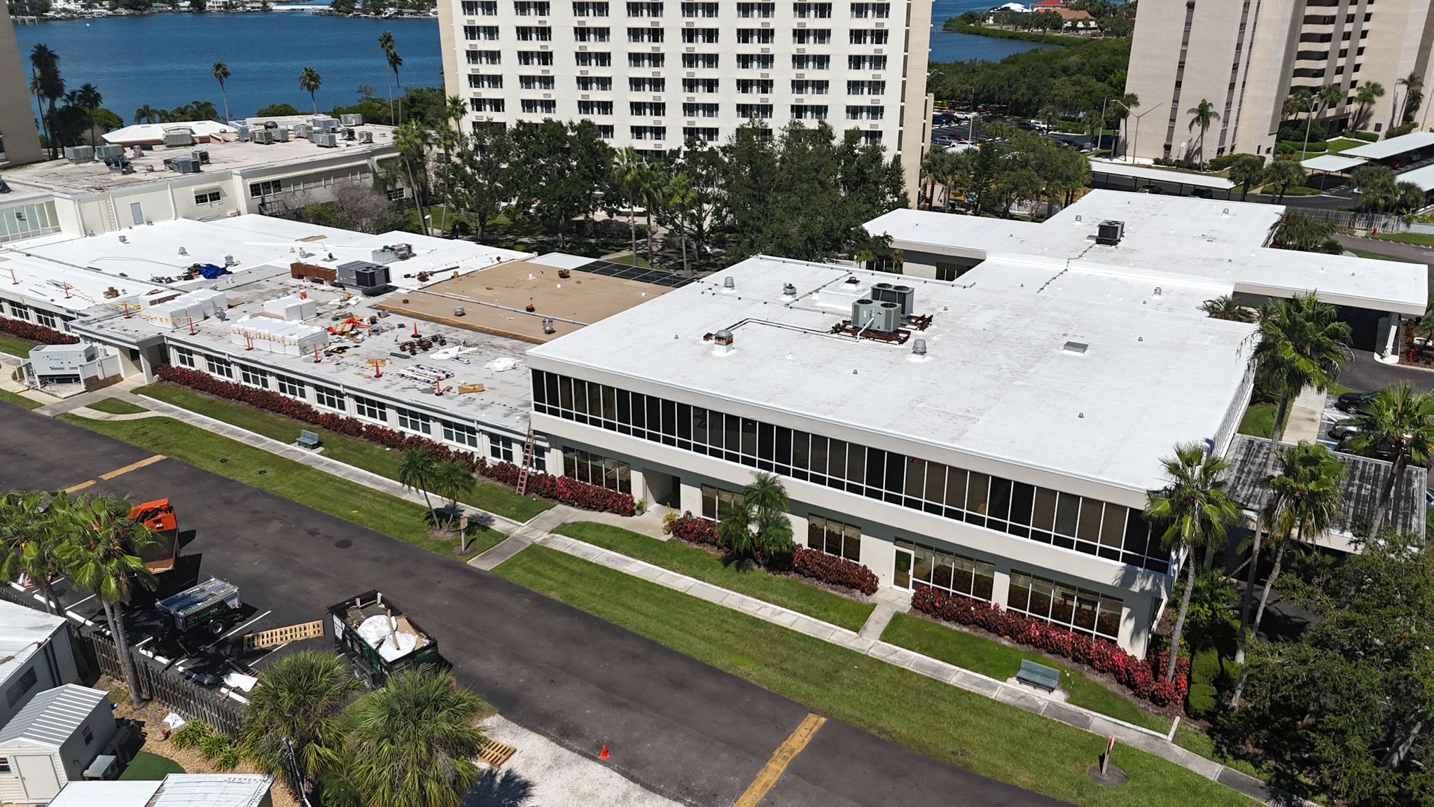 Aerial view of a white-roofed building complex with two main structures, surrounded by palm trees and grass, and tall apartment buildings and water visible in the background.