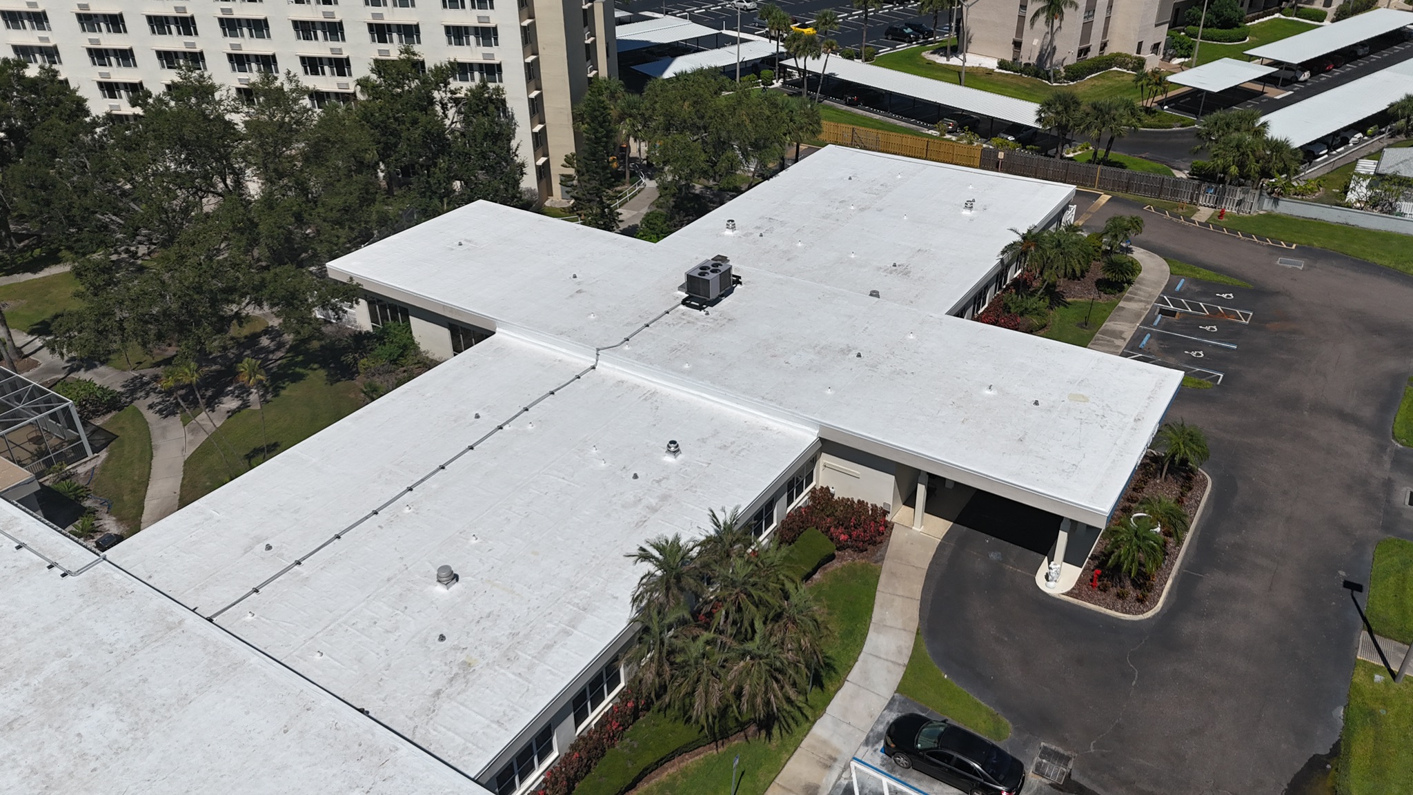 Aerial view of a white, flat-roofed building in a cross shape, surrounded by trees, landscaped greenery, a driveway, and parking spaces. Nearby, there are multi-story residential buildings and covered parking areas.